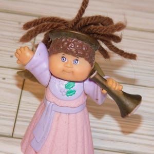 May include: A vintage Cabbage Patch Kids angel figurine. The doll has brown yarn hair, blue eyes, and is wearing a pink dress with a lavender sash and sleeves. It is holding a gold-colored trumpet. The figurine has the initials "CPK" on the chest.