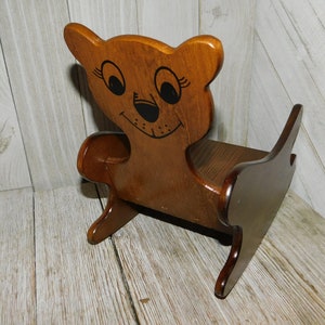 Puede incluir: Una silla mecedora de madera con forma de oso con la cara pintada. La silla es marr&oacute;n y tiene una cara pintada de negro con ojos grandes y una sonrisa.