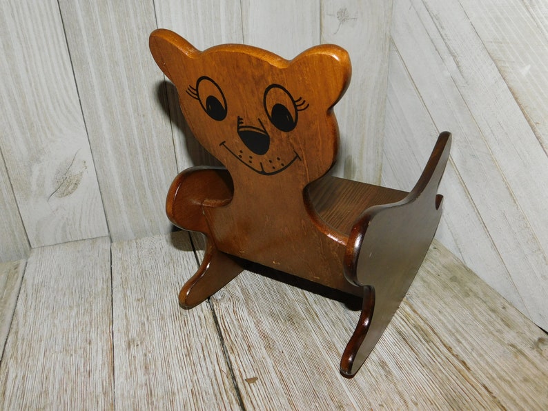 Puede incluir: Una silla mecedora de madera con forma de oso con la cara pintada. La silla es marr&oacute;n y tiene una cara pintada de negro con ojos grandes y una sonrisa.