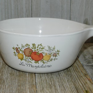 May include: White casserole dish with a handle, featuring a colorful illustration of tomatoes, lemons, and herbs. The words "La Marjolaine" are printed below the illustration. The dish is made of ceramic.
