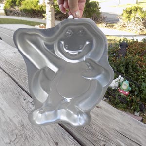 May include: A silver-colored, dinosaur-shaped cake pan. The pan is held up by a hand and is made of a shiny metal. The dinosaur has a smiling face and outstretched arms.