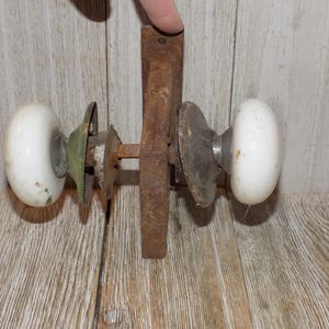 May include: Antique door hardware featuring two white oval doorknobs, a rusty metal plate, and various metal components. The hardware is mounted on a wooden surface, showcasing its vintage aesthetic.