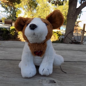 May include: A brown and white plush dog toy with a red heart on its chest. The dog is sitting on a wooden surface.