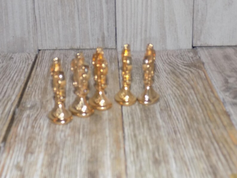 May include: Six gold-coloured chess pieces, including a king, queen, and rook, are arranged in a row on a wooden surface.