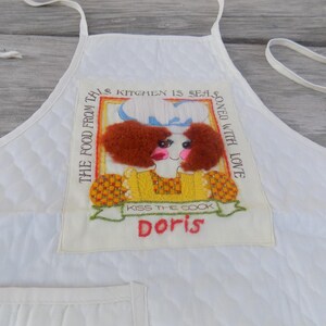 Op de afbeelding: Witte gewatteerde schort met een kleurrijk geborduurd ontwerp met een vrouw met rood haar en de tekst "The food from this kitchen is seasoned with love. Kiss the cook Doris."