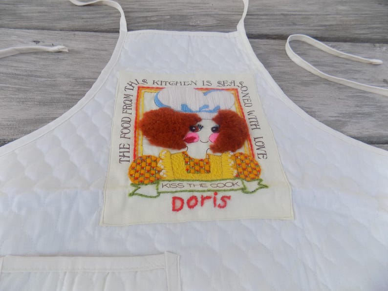 Op de afbeelding: Witte gewatteerde schort met een kleurrijk geborduurd ontwerp met een vrouw met rood haar en de tekst "The food from this kitchen is seasoned with love. Kiss the cook Doris."
