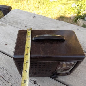Puede incluir: Una radio marr&oacute;n vintage con un asa negra y un dial. La radio mide aproximadamente 20 cm de alto, medido con una cinta m&eacute;trica amarilla. La radio tiene una rejilla de altavoz y un dial con n&uacute;meros.