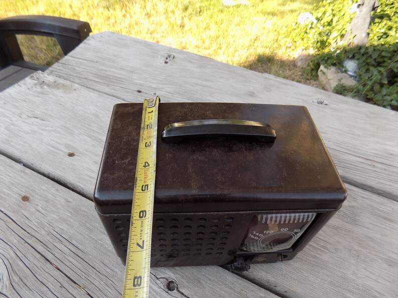 Puede incluir: Una radio marr&oacute;n vintage con un asa negra y un dial. La radio mide aproximadamente 20 cm de alto, medido con una cinta m&eacute;trica amarilla. La radio tiene una rejilla de altavoz y un dial con n&uacute;meros.