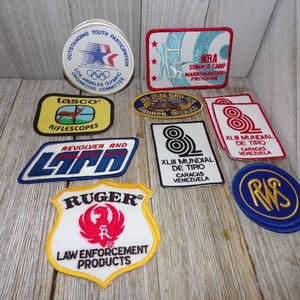 May include: A collection of vintage embroidered patches featuring various logos and designs, including "tasco riflescopes", "NRA Summer Camp Marksmanship Program", "Ruger Law Enforcement Products", and "XLIII Mundial de Tiro Caracas Venezuela".
