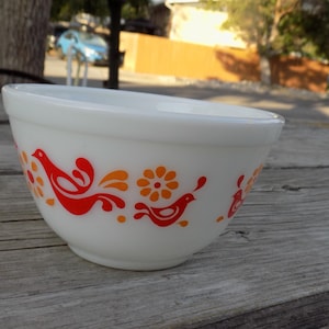 May include: A white milk glass mixing bowl featuring a repeating pattern of stylized orange and red birds and flowers around the exterior. The bowl has a smooth, rounded rim and a tapered base, showcasing a vibrant, colorful graphic design.