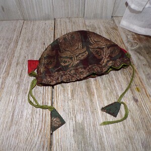 May include: A small, brown fabric pouch with a gold and green floral pattern. The pouch has a drawstring closure and green tassels.
