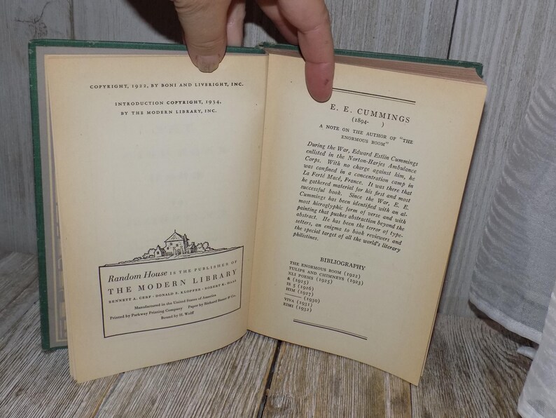 May include: A vintage book with a green cover titled "The Enormous Room" by E.E. Cummings. The book is open to a page with a note on the author and a bibliography of their works.