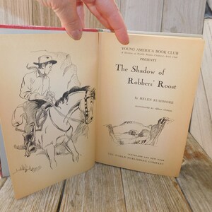 May include: A vintage book cover with a black and white illustration of a cowboy riding a horse. The title of the book is "The Shadow of Robbers' Roost" by Helen Rushmore. The book is published by The World Publishing Company.