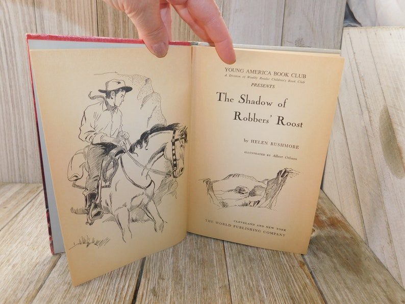 May include: A vintage book cover with a black and white illustration of a cowboy riding a horse. The title of the book is "The Shadow of Robbers' Roost" by Helen Rushmore. The book is published by The World Publishing Company.