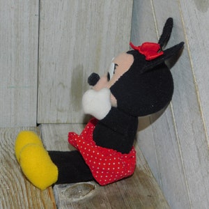May include: A black and white plush toy of Minnie Mouse wearing a red dress with white polka dots and a red bow. The toy is sitting on a wooden surface.