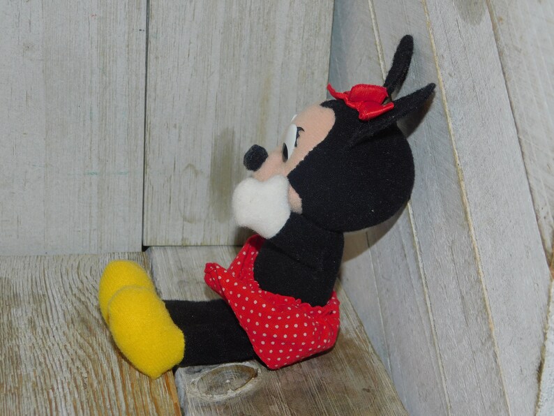 May include: A black and white plush toy of Minnie Mouse wearing a red dress with white polka dots and a red bow. The toy is sitting on a wooden surface.