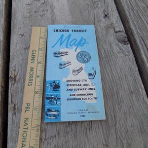 May include: A vintage Chicago Transit Map from 1954, featuring a light blue cover with illustrations of public transit vehicles. The map, approximately 3 inches long, is next to a wooden ruler. The text on the map reads "Chicago Transit Map".