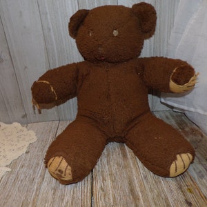 May include: A brown teddy bear with a worn, fuzzy texture. The bear has a stitched nose and brown button eyes. The bear's paws are patched with a tan fabric.