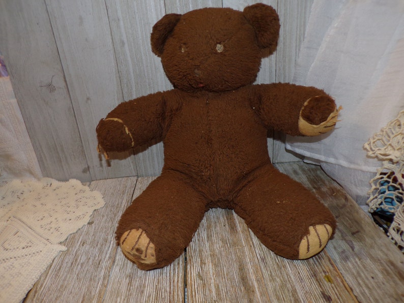 May include: A brown teddy bear with a worn, fuzzy texture. The bear has a stitched nose and brown button eyes. The bear's paws are patched with a tan fabric.