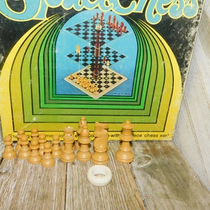 May include: Vintage Space Chess game set with a three-tiered board design. The box features a retro color scheme with the game's title in a stylized font. The chess pieces are light brown, and a white ring is also visible.