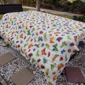 May include: A white tablecloth with a colorful butterfly and insect print. The tablecloth is spread out on a table in an outdoor setting.