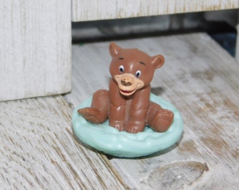 May include: A brown bear figurine sitting on a light blue base. The bear has a smiling expression with blue eyes and a light brown nose. The figurine is on a wooden surface.