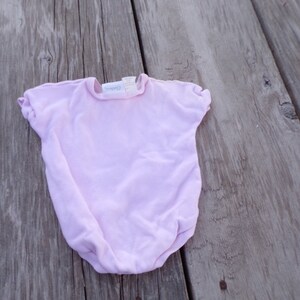May include: A pale pink baby onesie with short sleeves and a round neckline. The onesie has a small white tag with the word "Carter's" printed on it.