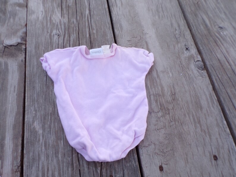 May include: A pale pink baby onesie with short sleeves and a round neckline. The onesie has a small white tag with the word "Carter's" printed on it.