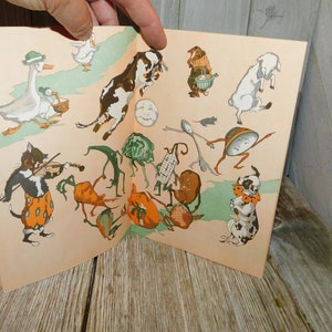 Op de afbeelding: Een vintage kinderboek met kleurrijke illustraties van dieren en groenten. Het boek is open op een pagina met een kat die viool speelt, een koe die over een maan springt, een gans die loopt, een hond met een bot en een verscheidenheid aan groenten met benen.