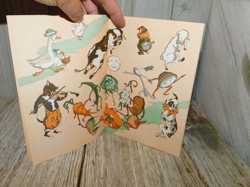 Op de afbeelding: Een vintage kinderboek met kleurrijke illustraties van dieren en groenten. Het boek is open op een pagina met een kat die viool speelt, een koe die over een maan springt, een gans die loopt, een hond met een bot en een verscheidenheid aan groenten met benen.