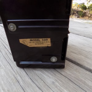 Puede incluir: Un dispositivo electr&oacute;nico vintage de color marr&oacute;n oscuro con forma rectangular. Una etiqueta en el lateral dice "MODELO 509" e incluye especificaciones el&eacute;ctricas. El dispositivo est&aacute; asegurado con sujetadores met&aacute;licos.