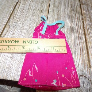 May include: A pink sleeveless dress with a floral print and a light blue ribbon tie at the top. The dress is approximately 6 inches long.