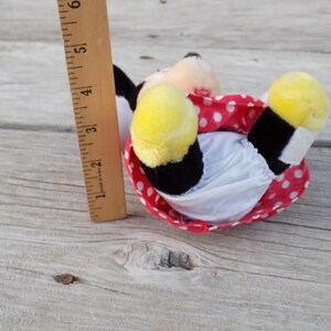 May include: A plush Minnie Mouse doll, lying on its back, with yellow feet, a red polka-dot skirt, and a white diaper. A wooden ruler, marked in inches, is next to the doll. The doll is approximately 5 inches long.