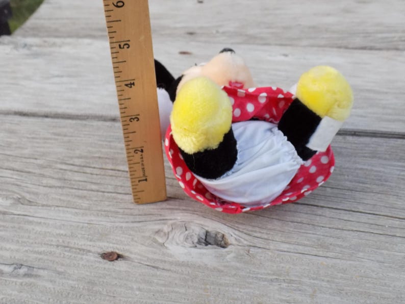 May include: A plush Minnie Mouse doll, lying on its back, with yellow feet, a red polka-dot skirt, and a white diaper. A wooden ruler, marked in inches, is next to the doll. The doll is approximately 5 inches long.