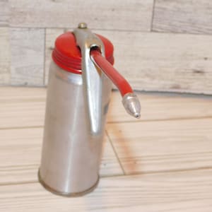 May include: A vintage metal oil can with a red top and a red spout. The can is silver-colored with a silver handle and spout tip. The oil can is likely used for lubricating machinery or tools.