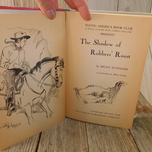 May include: An open book titled "The Shadow of Robbers' Roost" by Helen Rushmore, illustrated by Albert Orbaan. The book is published by The World Publishing Company in Cleveland and New York. The cover features a black and white illustration of a cowboy riding a horse.