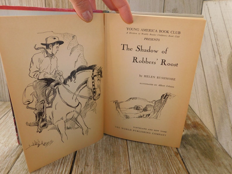 May include: An open book titled "The Shadow of Robbers' Roost" by Helen Rushmore, illustrated by Albert Orbaan. The book is published by The World Publishing Company in Cleveland and New York. The cover features a black and white illustration of a cowboy riding a horse.