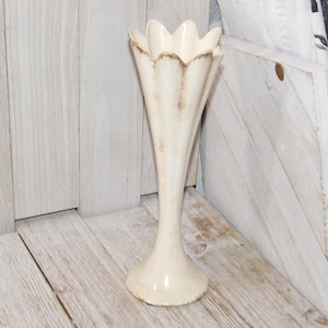 May include: A white ceramic vase with a fluted design and a scalloped rim. The vase has a tall, slender shape and a wide base.