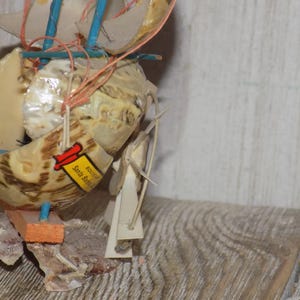 May include: A handcrafted seashell sailboat with tan and brown shells, blue and orange accents, and a small tag that reads "Santa Barbara". The miniature boat features multiple sails and is assembled with string.