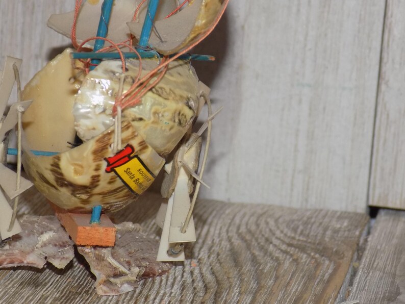 May include: A handcrafted seashell sailboat with tan and brown shells, blue and orange accents, and a small tag that reads "Santa Barbara". The miniature boat features multiple sails and is assembled with string.