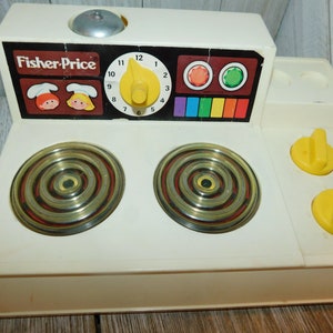 May include: A vintage Fisher-Price toy stove in off-white plastic featuring two circular burners with concentric rings, a central yellow timer knob, two yellow control dials, and a control panel with colorful graphics and two cartoon chef faces.