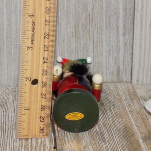 May include: A wooden nutcracker figurine with a red, blue, and green costume. The nutcracker is holding two white balls in its hands. The nutcracker is standing on a green base with the words "Made in China" printed on it.