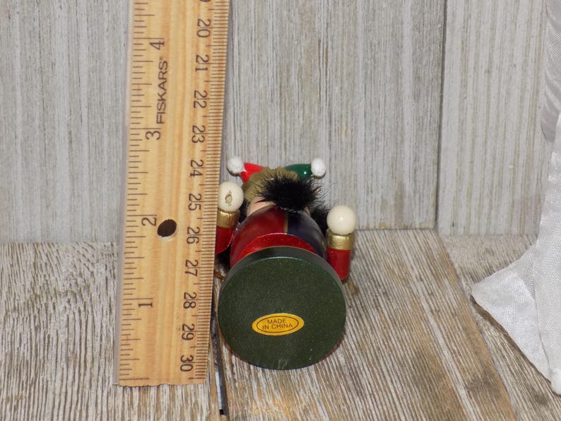 May include: A wooden nutcracker figurine with a red, blue, and green costume. The nutcracker is holding two white balls in its hands. The nutcracker is standing on a green base with the words "Made in China" printed on it.