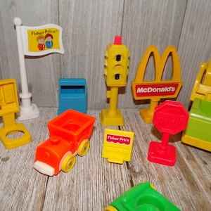 May include: A collection of vintage Fisher-Price Little People playset pieces, including a yellow train, a red stop sign, a yellow traffic light, a blue mailbox, a yellow phone booth, a yellow McDonald's sign, and a yellow and green construction vehicle.