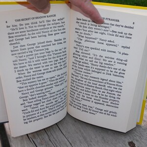 May include: An open book with yellow pages and a yellow spine. The book is titled "The Secret of Shadow Ranch" and "The Stranger". The text on the pages describes a ghostly horse and a romantic legend about a local sheriff.