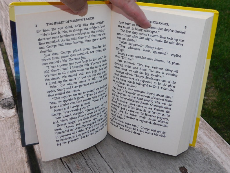 May include: An open book with yellow pages and a yellow spine. The book is titled "The Secret of Shadow Ranch" and "The Stranger". The text on the pages describes a ghostly horse and a romantic legend about a local sheriff.