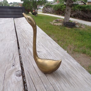 Pu&ograve; includere: Una scultura di cigno in metallo dorato con un collo lungo e un corpo curvo. Il cigno &egrave; seduto su una superficie di legno.