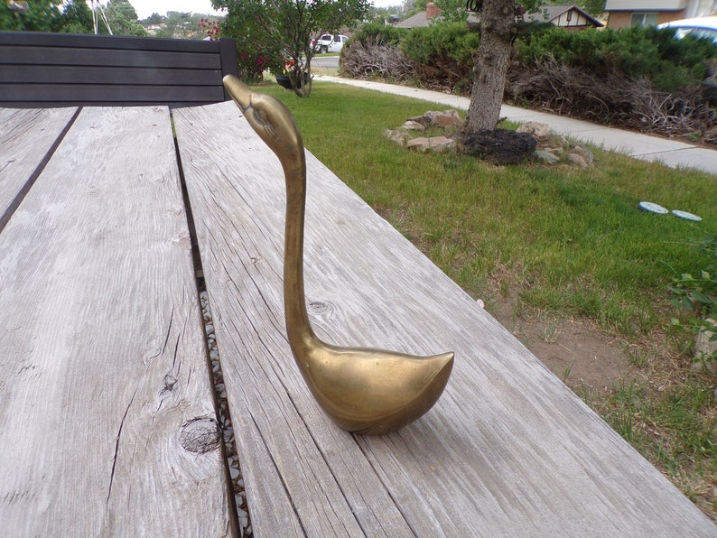 Pu&ograve; includere: Una scultura di cigno in metallo dorato con un collo lungo e un corpo curvo. Il cigno &egrave; seduto su una superficie di legno.