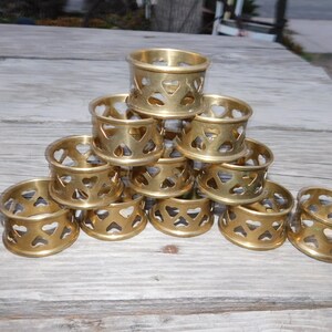 May include: A collection of gold-colored napkin rings. Each ring features heart-shaped cutouts around the band. Some rings are stacked in a pyramid shape, while others are scattered on a wooden surface.