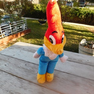 May include: A plush toy of Woody Woodpecker, a cartoon character. The toy is red, yellow, and blue. It is standing on a wooden surface.